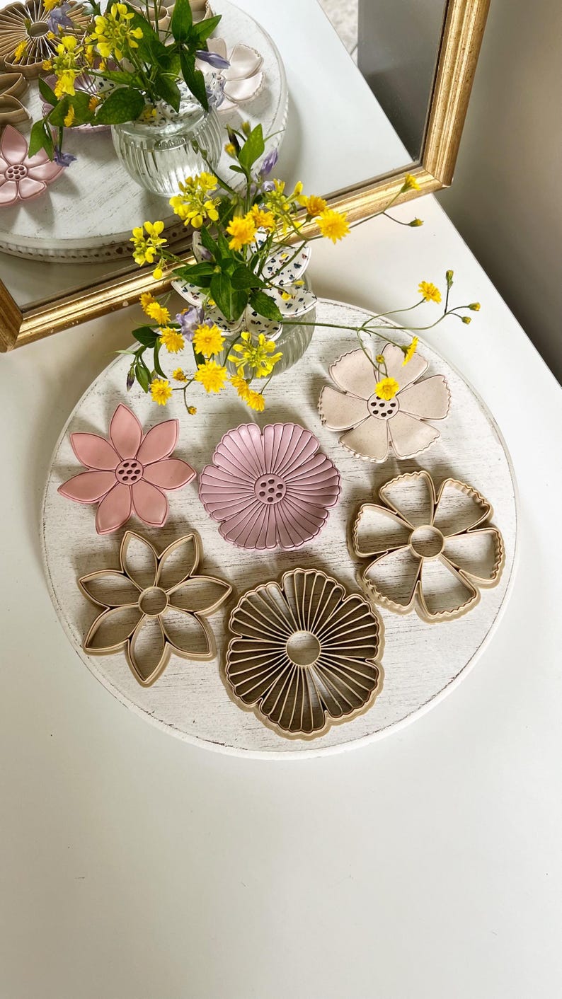 May include: A collection of flower-shaped items in various colors, including pink and beige, arranged on a white, round surface. The items are displayed with a vase of yellow flowers and a mirror in the background. The items appear to be decorative or for crafting.