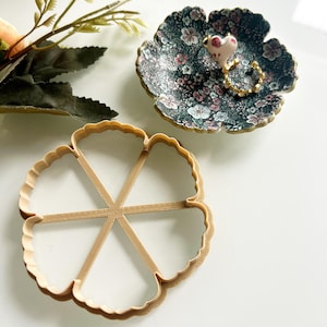 May include: A floral-patterned trinket dish with a small frog figurine and a gold ring. A wooden flower-shaped cookie cutter is also present. Decorative sprigs of greenery are in the background. The items are on a white surface.