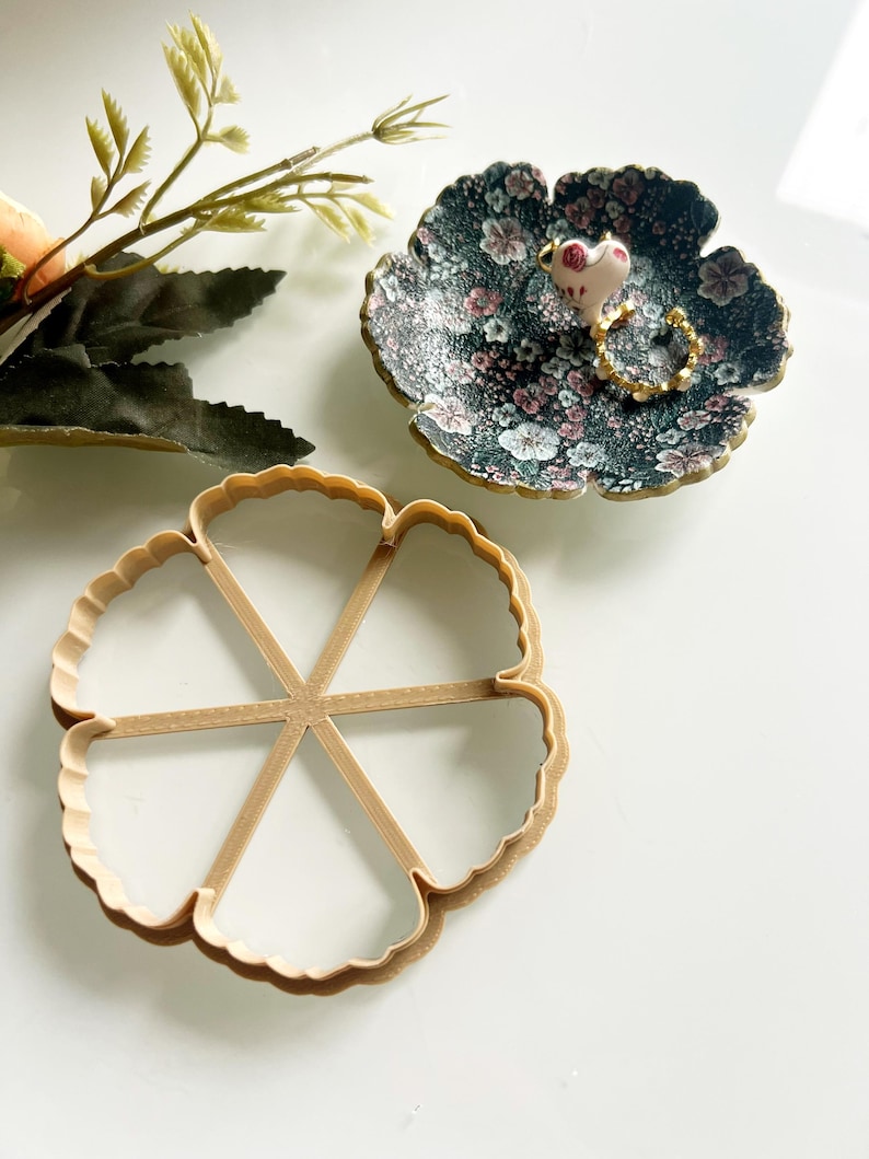 May include: A floral-patterned trinket dish with a small frog figurine and a gold ring. A wooden flower-shaped cookie cutter is also present. Decorative sprigs of greenery are in the background. The items are on a white surface.