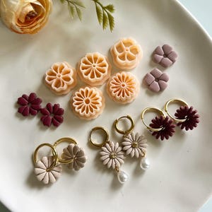 Puede incluir: Una colección de pendientes florales hechos a mano y objetos decorativos con forma de flor en varios colores, incluyendo melocotón, burdeos y rosa claro. Algunos pendientes tienen aros dorados y detalles de perlas. Los artículos están dispuestos en un plato ovalado blanco.