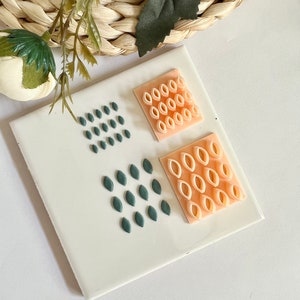 Puede incluir: Un azulejo de cerámica blanco con tres diseños geométricos. Los diseños están hechos de pequeñas formas en relieve. El diseño superior es un cuadrado hecho de óvalos de color melocotón. El diseño izquierdo es un cuadrado hecho de óvalos verde oscuro. El diseño derecho es un cuadrado hecho de óvalos de color melocotón.