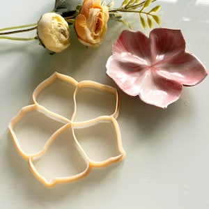 Puede incluir: Un cortador de galletas de plástico beige con forma de flor de cinco pétalos redondeados y un plato de cerámica rosa a juego con forma de flor y veteado blanco, acompañado de dos rosas artificiales de color crema y melocotón con tallos y hojas.
