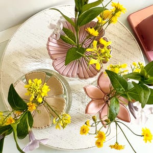 May include: Three flower-shaped dishes in shades of pink and beige, each holding a small arrangement of yellow flowers and green leaves. The dishes sit on a white, round, distressed wooden surface.