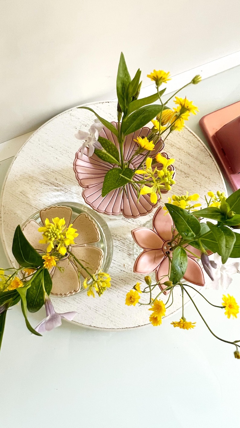 May include: Three flower-shaped dishes in shades of pink and beige, each holding a small arrangement of yellow flowers and green leaves. The dishes sit on a white, round, distressed wooden surface.