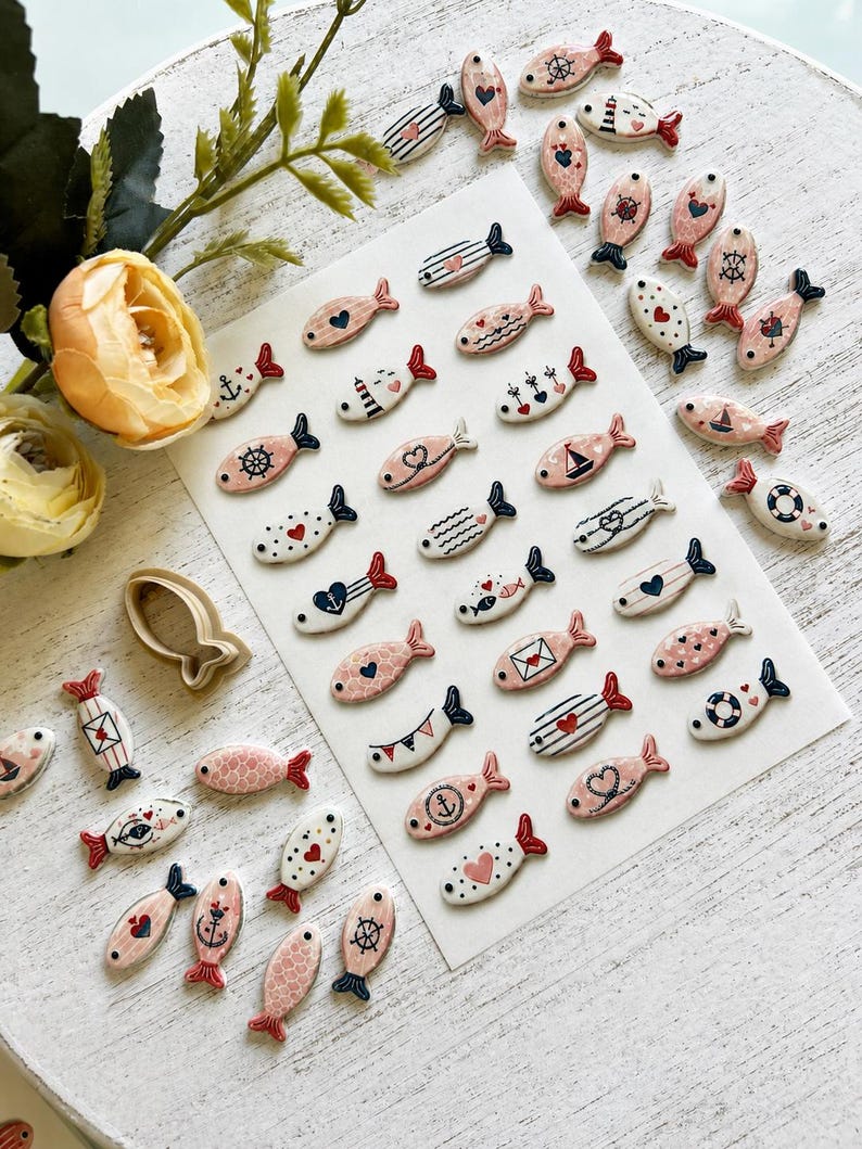 May include: A collection of small, flat, fish-shaped ceramic or clay pieces featuring various nautical and heart-themed patterns in pink, white, navy blue, and red. The set includes a single fish-shaped cookie cutter and loose pieces.