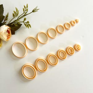 Könnte beinhalten: Eine Sammlung oval geformter Ausstecher in verschiedenen Größen, angeordnet in zwei Reihen. Die Ausstecher sind hellpfirsichfarben und scheinen aus Kunststoff zu sein. Ein Zweig mit Grün und eine Blume befinden sich in der oberen linken Ecke.