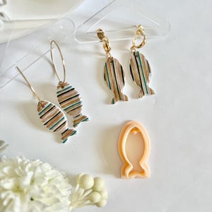 Puede incluir: Un conjunto de tres pendientes con forma de pez con un patrón a rayas en tonos marrones, blancos y azules. Los pendientes están hechos de arcilla polimérica y tienen ganchos dorados. También se incluye un cortador de galletas con forma de pez de plástico.