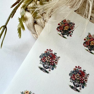 May include: A sheet of white paper with multiple floral bouquet stickers. The stickers feature a variety of flowers in shades of red, yellow, blue, and white, arranged in a decorative vase. The background includes woven textures and greenery.