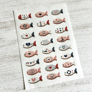 May include: A collection of small, flat, fish-shaped decorative pieces arranged in a grid on a white sheet. Each fish features unique nautical and romantic patterns in shades of red, navy blue, and soft pink, including anchors, hearts, stripes, and sailboats.