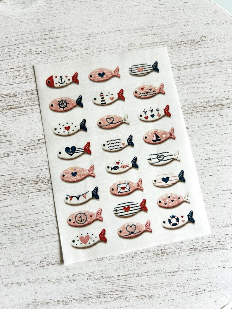 May include: A collection of small, flat, fish-shaped decorative pieces arranged in a grid on a white sheet. Each fish features unique nautical and romantic patterns in shades of red, navy blue, and soft pink, including anchors, hearts, stripes, and sailboats.