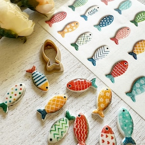 Può includere: Una collezione di piccoli ornamenti in ceramica a forma di pesce con vari motivi come strisce, pois e chevron, insieme a un tagliabiscotti in plastica color marrone chiaro e un foglio di adesivi a forma di pesce abbinati.
