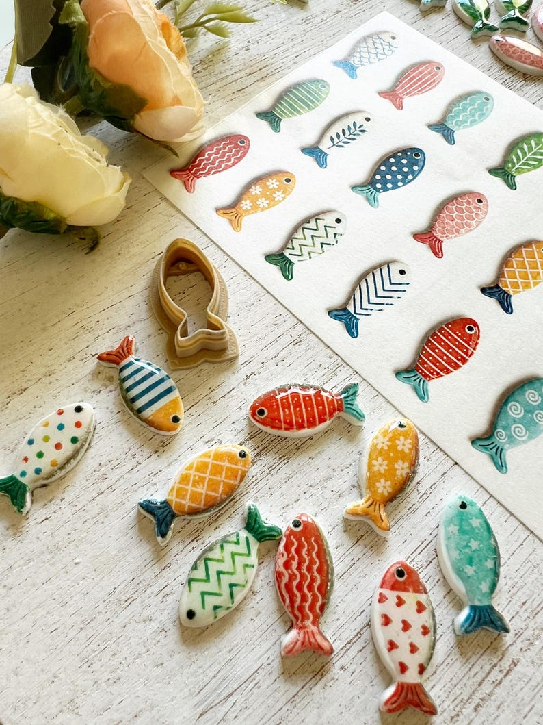 May include: A collection of small, colorful ceramic fish-shaped ornaments featuring various patterns like stripes, dots, and chevrons, alongside a single tan plastic fish-shaped cookie cutter and a sheet of matching fish stickers.