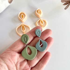Puede incluir: Dos pares de pendientes en forma de lágrima en verde salvia y azul apagado, cada uno con un pequeño pendiente superior y una pieza inferior colgante. Detrás hay dos cortadores de plástico color melocotón para dar forma a los diseños.