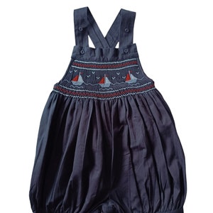 Peut inclure: Une salopette bébé bleu foncé smockée avec un motif de voilier blanc et rouge. La salopette a des bretelles réglables avec des boutons.