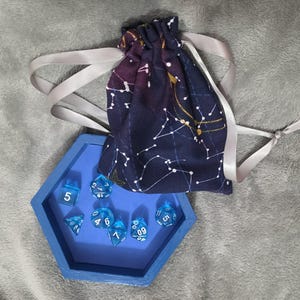 Puede incluir: Una bandeja de madera azul contiene un juego de dados poliédricos azules transparentes. Una bolsa con cordón con un patrón de constelaciones celestes en blanco y dorado está al lado de la bandeja. La bolsa tiene un fondo azul oscuro con acentos morados y una cinta plateada.