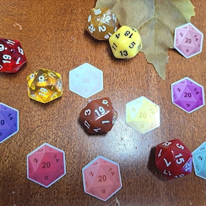 May include: A collection of 20-sided dice in various colors, including red, yellow, purple, and brown. The dice are arranged on a wooden surface with stickers of dice with different numbers.