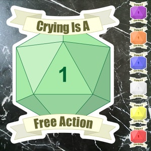 May include: A green 20-sided die with the number 1 on it. The die is surrounded by a white banner with the text "Crying Is A Free Action".