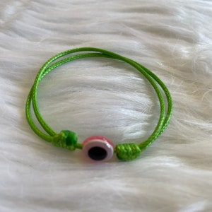 May include: A green cord bracelet featuring a pink and white eye-shaped bead with a black center. The bracelet is knotted at each end and is displayed on a white, fluffy surface. The bracelet is a simple, yet stylish accessory.