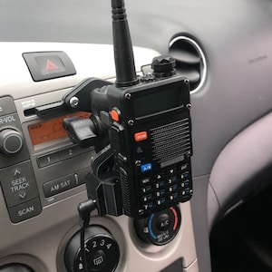 May include: A black Baofeng radio mounted in a car. The radio has a screen, buttons, and an antenna. The mount is attached to the car's dashboard. The radio is black with orange and blue accents.