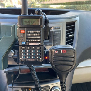 May include: A black and gray two-way radio with the display showing "NOAA 2 146.640". The radio is mounted in a car, next to a black BTECH microphone. The radio has multiple buttons and a small antenna.