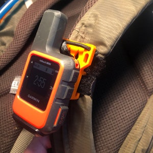 May include: An orange and black Garmin inReach Mini GPS device attached to a backpack strap with an orange clip. The device displays the time 2:55 and the date Sat 29. The backpack is brown and green.