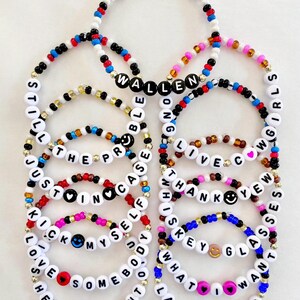 May include: A collection of beaded bracelets in various colors, featuring white letter beads spelling out phrases like "WALLEN," "KICK MYSELF," and "WHAT I WANT." The bracelets include smiley faces and heart-shaped beads. The text "BRACELETS ARE SOLD INDIVIDUALLY" is at the bottom.