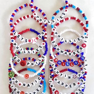 May include: A collection of beaded bracelets in red, white, and blue. Some bracelets spell out "PHILLIES", "LET'S GO", and player names. Smiley faces and baseballs are also featured. The text "BRACELETS ARE SOLD INDIVIDUALLY" is at the bottom.