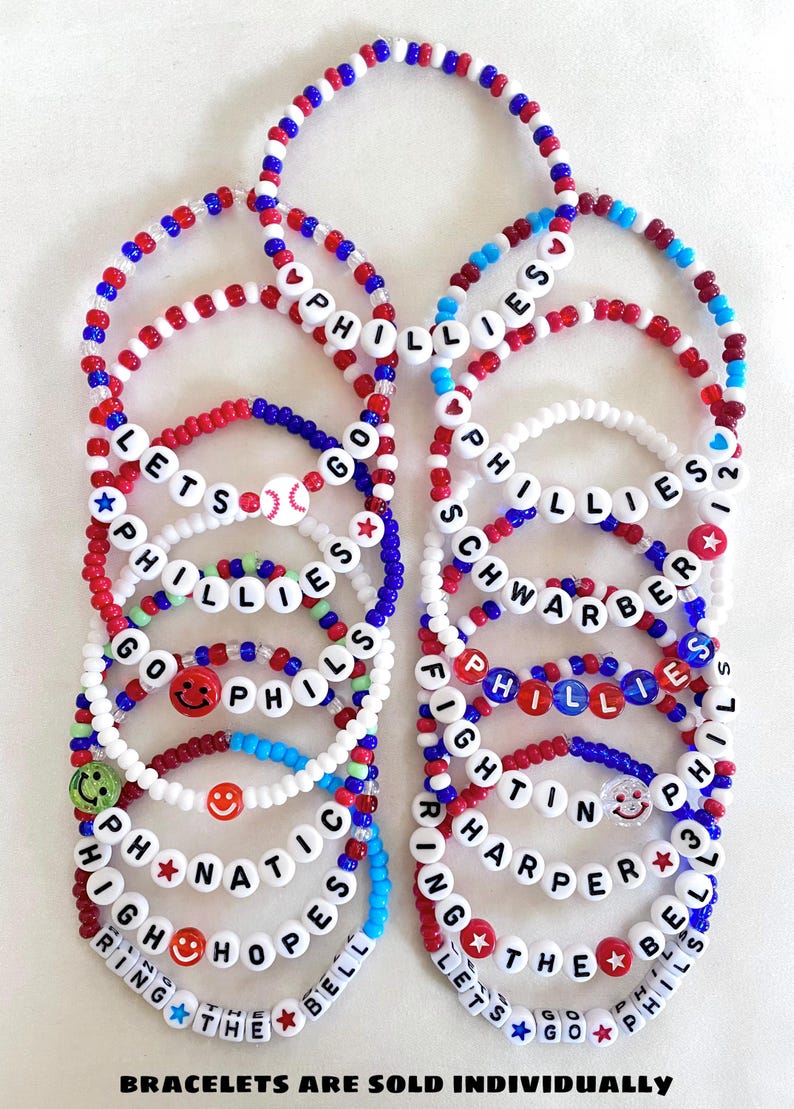May include: A collection of beaded bracelets in red, white, and blue. Some bracelets spell out "PHILLIES", "LET'S GO", and player names. Smiley faces and baseballs are also featured. The text "BRACELETS ARE SOLD INDIVIDUALLY" is at the bottom.