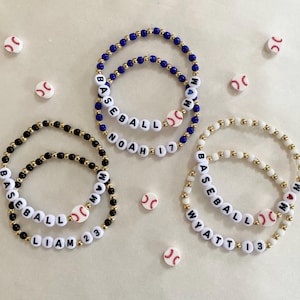 May include: Three beaded bracelets with baseball-themed charms. The bracelets are black, white, and blue with gold beads. The bracelets spell out the names "Liam", "Wyatt", and "Noah".