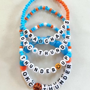 May include: Four beaded bracelets in blue, orange, and white. The bracelets spell out "OKC THUNDER" and "NBA CHAMPS" with star and basketball bead accents. The bracelets are sold individually.