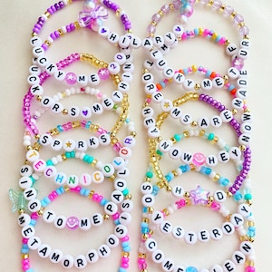 May include: A collection of colorful beaded bracelets featuring white letter beads that spell out phrases like Lucky Me, Technicolor, Metamorphosis, Why Not, Dreams Are, Hey Now, So Yesterday, Come Clean, and With Love, accented with stars and hearts.