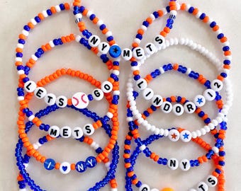 May include: A collection of beaded bracelets in blue, orange, and white. Some bracelets spell out "NY METS", "LET'S GO METS", and "LINDOR 12". Other bracelets have a baseball, star, and smiley face beads. The text "BRACELETS ARE SOLD INDIVIDUALLY" is at the bottom.