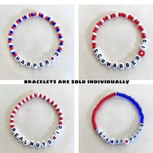 May include: Four beaded bracelets in red, white, and blue. Each bracelet features white letter beads spelling out names and phrases, with star accents. The text "BRACELETS ARE SOLD INDIVIDUALLY" is also present.