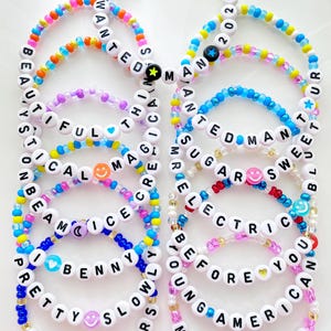 May include: A collection of colorful beaded bracelets with white letter beads spelling out words like "BEAUTIFUL", "WANTED", and "AMERICA". Each bracelet features a unique color scheme and decorative beads. The text "BRACELETS ARE SOLD INDIVIDUALLY" is at the bottom.