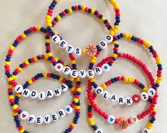 May include: A collection of stretch bracelets made with yellow, red, and dark blue beads. The bracelets feature white letter beads spelling LETS GO, FEVER, INDIANA, CLARK 22, BOSTON 7, and include basketball charms, stars, and hearts.