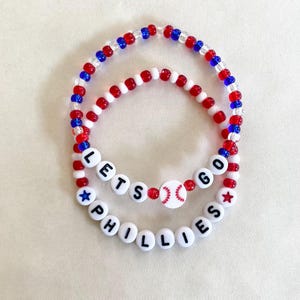 May include: Two beaded bracelets. The top bracelet has red, white, and blue beads. The bottom bracelet has white beads with black letters that spell "PHILLIES" and "LETS GO". A baseball bead is in the center.
