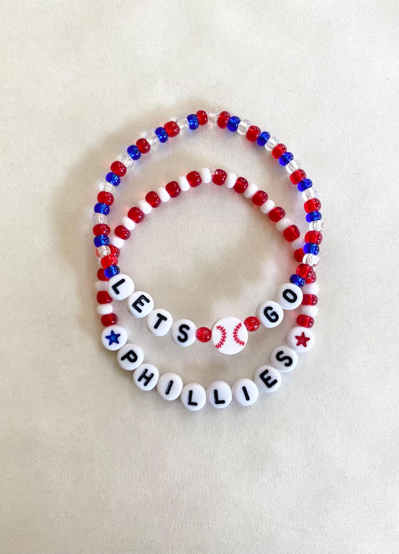 May include: Two beaded bracelets. The top bracelet has red, white, and blue beads. The bottom bracelet has white beads with black letters that spell "PHILLIES" and "LETS GO". A baseball bead is in the center.