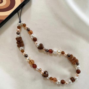 May include: A brown, white, and gold beaded phone strap with brown teddy bear charms and brown heart charms.