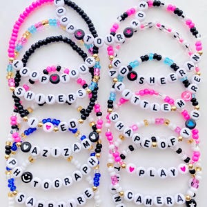 May include: A collection of beaded bracelets featuring white letter beads spelling out song titles and tour names like Loop Tour 2026, Ed Sheeran, Shivers, A Little More, Shape of You, Photograph, Sapphire, Perfect, Play, Camera, and Thinking Out Loud.