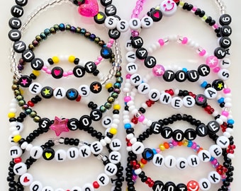 May include: A collection of beaded bracelets in various colors, featuring white letter beads spelling out words and names. Some bracelets include heart, star, and smiley face charms. The text "BRACELETS ARE SOLD INDIVIDUALLY" is at the bottom.