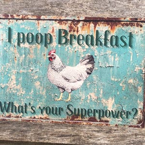 Pode incluir: Uma placa de metal enferrujada com fundo azul e uma ilustração de frango branco. A placa diz "Eu caco no café da manhã. Qual é o seu superpoder?"