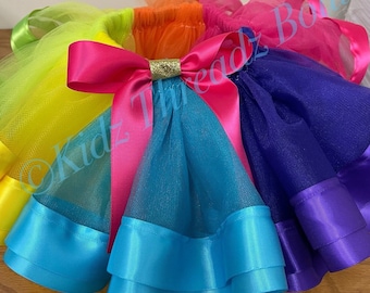 May include: A rainbow tulle skirt with a pink satin bow and gold glitter accent. The skirt has layers of turquoise, purple, and yellow tulle with matching satin ribbon trim.