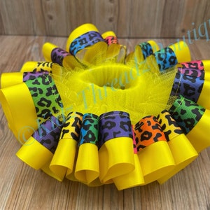 May include: A yellow tulle bow with a rainbow leopard print ribbon. The bow is made of multiple layers of tulle and ribbon, and is tied with a yellow ribbon.