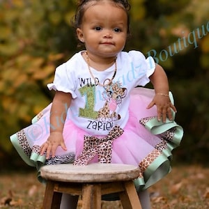 May include: A white short-sleeved t-shirt with a green glittery number one, a leopard cub graphic, and the text Wild 1 Zariel, paired with a layered pink tulle tutu skirt featuring leopard print and light green ribbon trim accents.