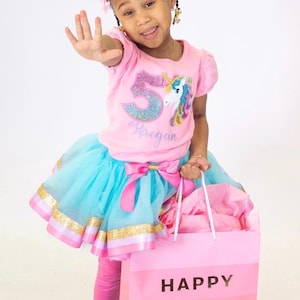 May include: A young girl wearing a pink t-shirt with a glittery number 5 and a unicorn graphic, paired with a blue tutu skirt and pink leggings. She holds a pink gift bag with "HAPPY BIRTHDAY" text.
