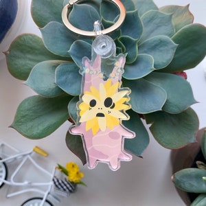 May include: A clear acrylic keychain featuring a cartoon character with a yellow sunflower head and pink body. The keychain has a silver metal ring. The background includes a succulent plant and small decorative items.