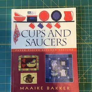Puede incluir: Un libro titulado "Tazas y platillos: Diseños de cocina con piezas de papel" de Maaike Bakker. La portada presenta un esquema de color rojo, blanco y azul con ilustraciones de tazas, platillos y una tetera.