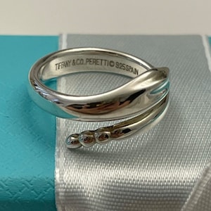 May include: A silver ring with a distinctive, flowing design. The ring has a wrap-around style with a textured band and small, round embellishments. The inscription "TIFFANY & CO. PERETTI © 925 SPAIN" is visible on the inside of the ring.