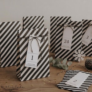 May include: A collection of black and white diagonal striped paper bags for an advent calendar. Each bag is tied with black and white twine and features a white rectangular tag with a printed number and the text NOCH 23 TAGE BIS WEIHNACHTEN.