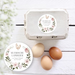 Könnte beinhalten: Ein weißer Eierkarton mit einem Aufkleber mit der Aufschrift "10 FRISCHE LANDEIER" auf Deutsch, umgeben von einem floralen Kranz und einer Hühnerillustration. Drei Eier liegen neben dem Karton. Die Eier sind hellbraun.